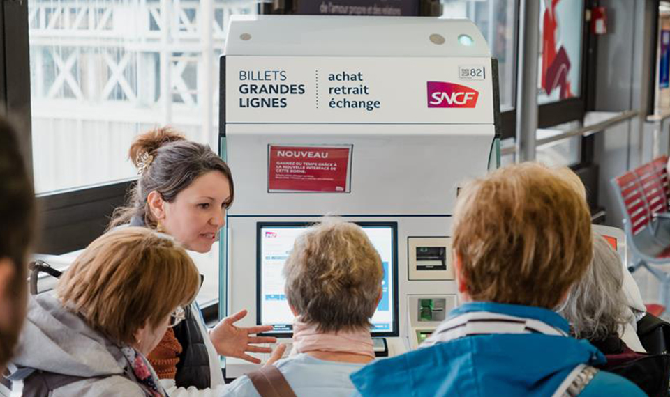 Une agente aide des voyageurs devant une borne SNCF